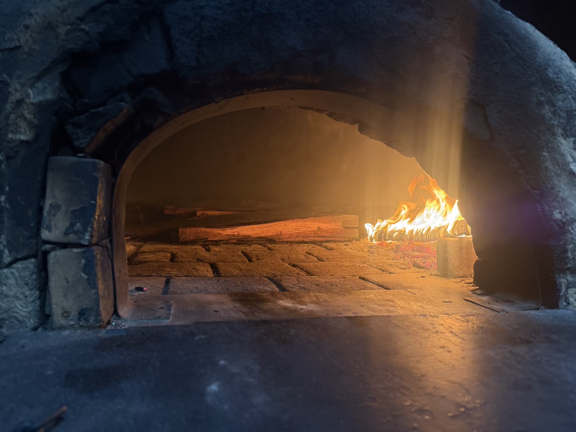 El horno de leña real de Mambo Italiano encendido, con la llama viva y la luz atravesando el humo dentro del arco de piedra